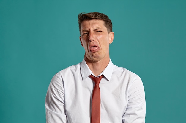 Portrait of an attractive brunet guy with brown eyes, wearing in a classic white shirt and red tie. He is acting like does not like something while posing in a studio against a blue background. Concep