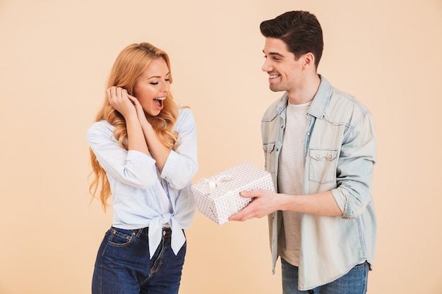 Portrait of attractive brunette man giving birthday box to beautiful young woman, isolated over beige wall