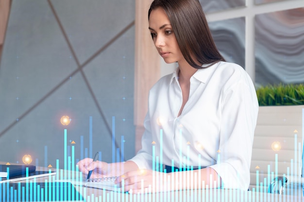 portrait of attractive businesswoman in formal wear working with documents and thinking how to optimize trading strategy at corporate finance fund forex chart hologram over modern office background