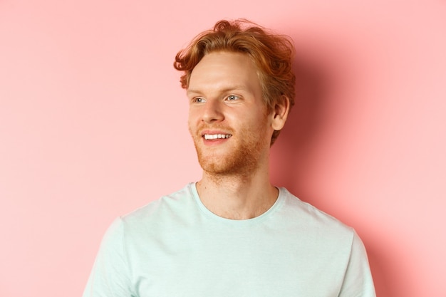 Portrait of attractive caucasian guy with red messy hair and beard, turn head and looking left with pleased smile, standing over pink background