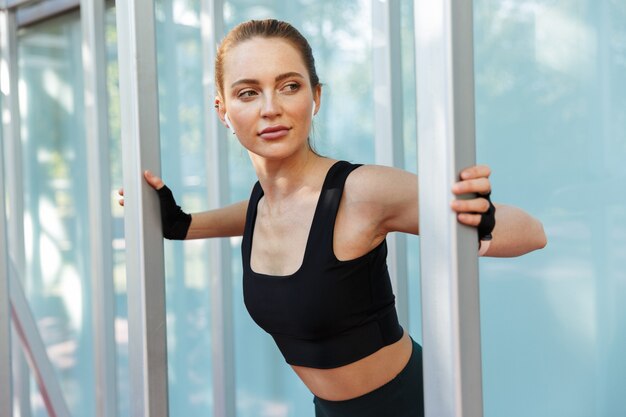 Portrait of attractive caucasian woman wearing tracksuit doing workout with horizontal metal bar on sports ground in green park