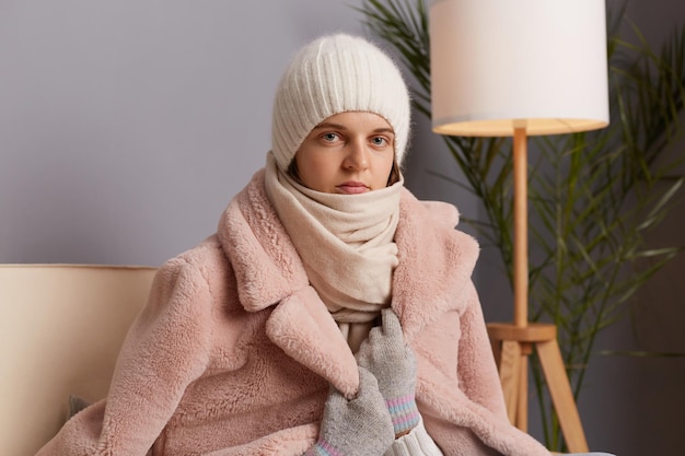 Portrait of attractive Caucasian young adult woman wearing warm clothes and wrapped in scarf sitting on sofa in room looking at camera feels icily being sad