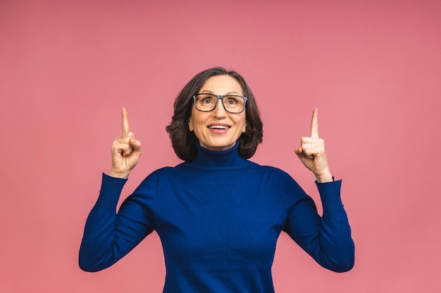 Portrait of attractive cheerful beautiful senior mature aged woman demonstrating copy space isolated over pink background, pointing finger up.