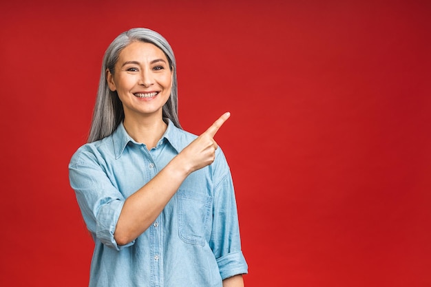 Portrait of attractive cheerful beautiful senior mature aged woman demonstrating copy space isolated over red background pointing finger away