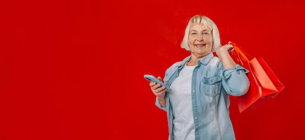 Portrait of attractive cheerful blonde 50s woman using device holding bags order delivery isolated over red pastel color background