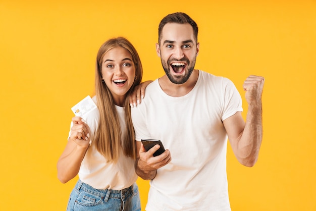 Portrait of an attractive cheerful young couple wearing casual clothing standing isolated over yellow wall, celebrating while using mobile phone and showing credit card