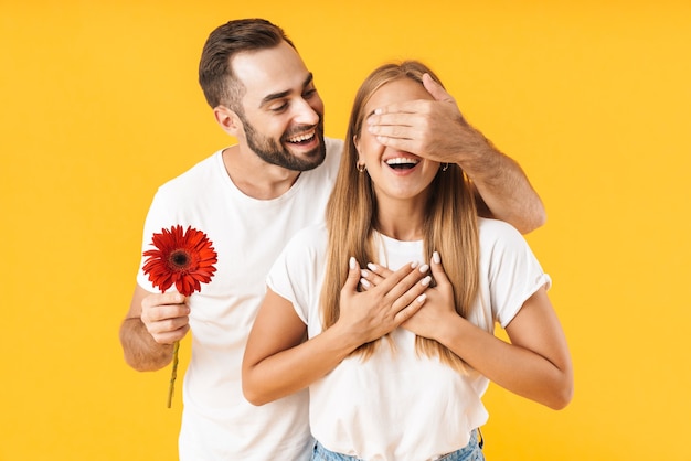 Portrait of an attractive cheerful young couple wearing casual clothing standing isolated over yellow wall, celebrating, woman is holding a flower