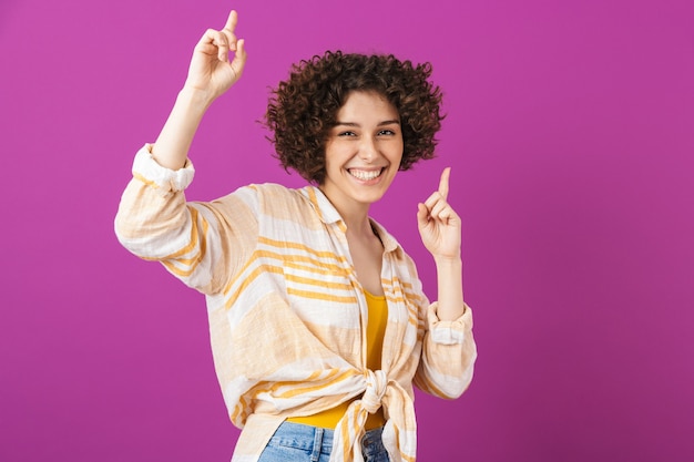 Portrait of an attractive cheerful young woman with curly brunette hair standing isolated over violet wall, celebrating success