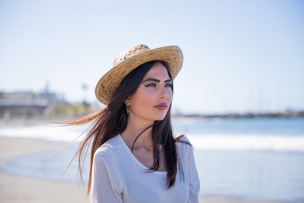 Portrait of an attractive and confident caucasian girl at the beach. She relaxes and wears a summer hat