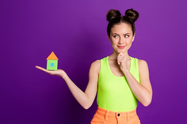 Portrait of attractive curious cheery girl holding on palm house figure thinking isolated on bright violet color background