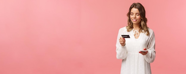 Portrait of attractive and cute blond girl with short curly hairstyle white dress holding smartphone and credit card insert digit numbers to register in shopping app buy online pink background