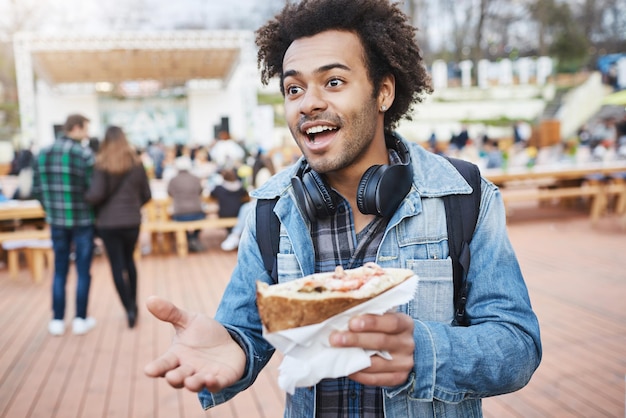 旅行中のフードフェスティバルでサンドイッチを食べている友人と何かを話している間、アフロヘアーを身振りで示す魅力的な浅黒い肌の男の肖像男はガールフレンドと食べ物を共有したい