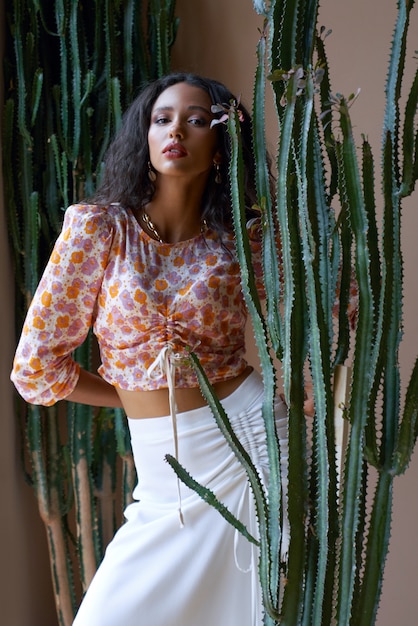 Ritratto di ragazza attraente mulatta alla moda con lunghi capelli neri ricci in posa tra i cactus alti al chiuso.
