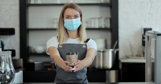 Portrait of attractive female barista in madical mask and holding beautiful coffee to go, barista coffee shop lifestyle, coffee creations.