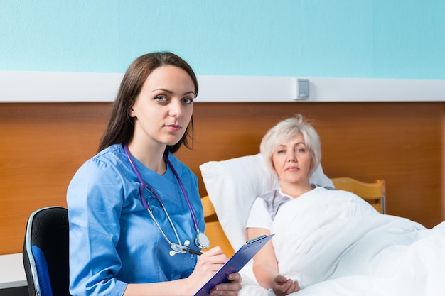 Foto ritratto di attraente dottoressa in uniforme che annota i reclami del paziente, che giace nel letto d'ospedale nel reparto ospedaliero. concetto di assistenza sanitaria