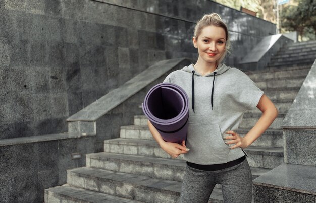 Donna attraente di forma fisica del ritratto con la stuoia arrotolata nelle sue mani all'aperto. uno stile di vita sano