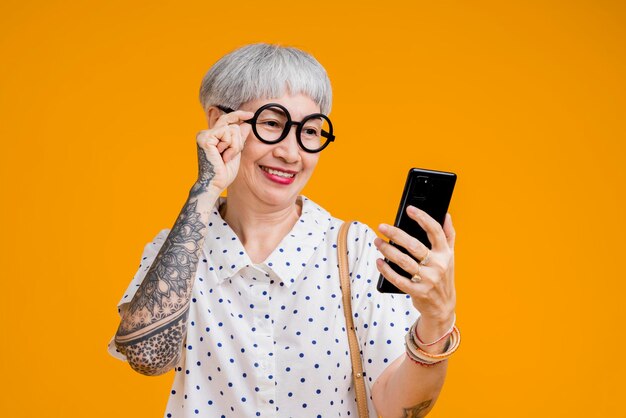 Portrait of attractive funky asian tattoo cheerful greyhaired elder old senior woman touching spectacles and smartphone device internet conected isolated over yellow pastel background color