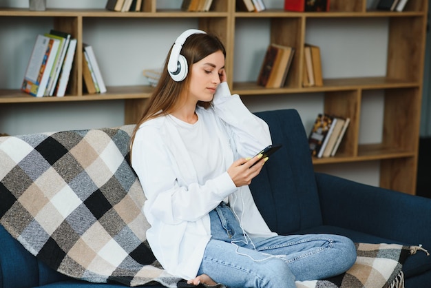 Portrait of attractive girl 20s wearing headphones using smartphone and listening to music while sitting on sofa at home