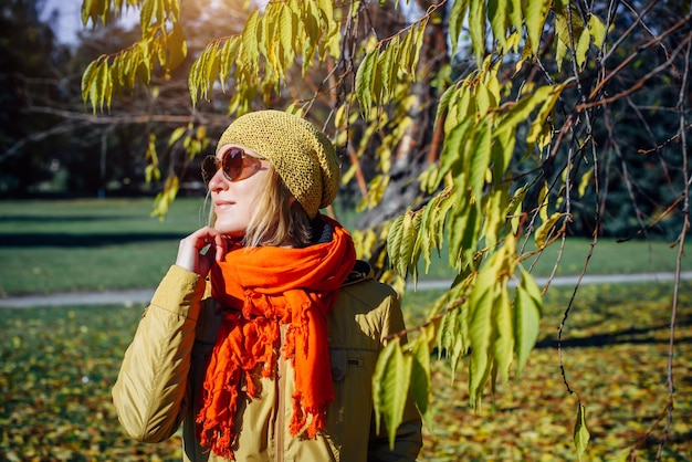 Portrait of attractive girl in sunglasses and red scarf on the background of yellow foliage in autumn park. Young woman enjoying walk on fall sunny day.
