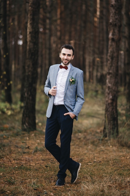 The portrait of attractive groom in a suit and bow tie with boutonniere or buttonhole on jacket, is standing against the background of the forest at nature.