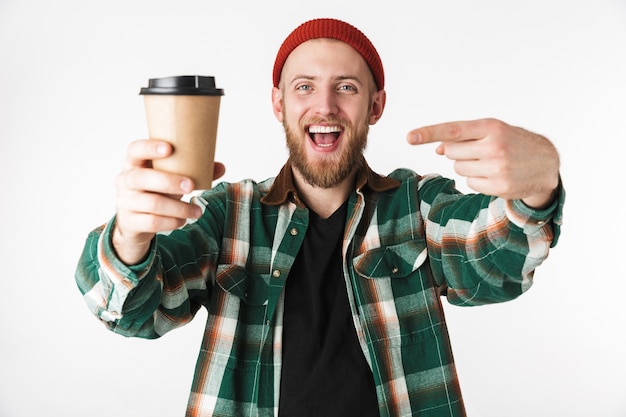 Portrait of attractive guy wearing hat and plaid shirt holding paper cup with coffee, while standing isolated over white background