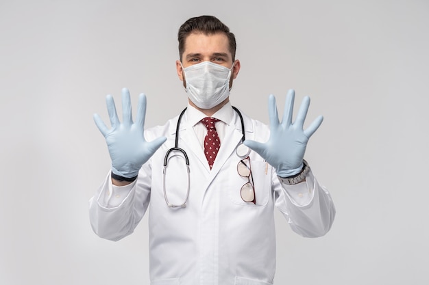 Portrait of attractive handsome doctor in protective face mask, white lab coat, tie isolated on white wall