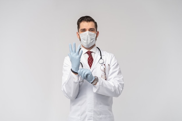 Portrait of attractive handsome doctor in protective face mask, white lab coat, tie isolated on white wall