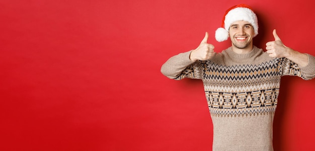 Portrait of attractive happy man in santa hat and sweater showing thumbsup in approval and smiling w...