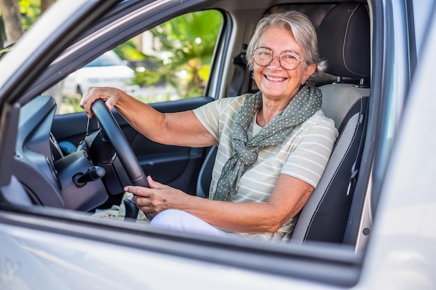 手で彼女の車に座っている t シャツの魅力的な幸せな年配の女性の肖像画はカメラの笑顔を見てステアリング ホイールを保持します