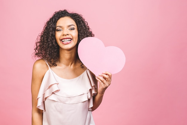 Портрет привлекательной счастливой улыбающейся американской афро женщины изолированной на розовом фоне с большим розовым сердцем.