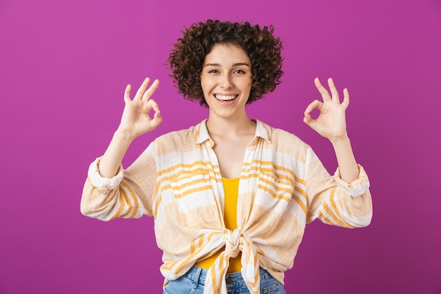 Portrait of an attractive happy young woman with curly brunette hair standing isolated over violet wall, showing ok gesture