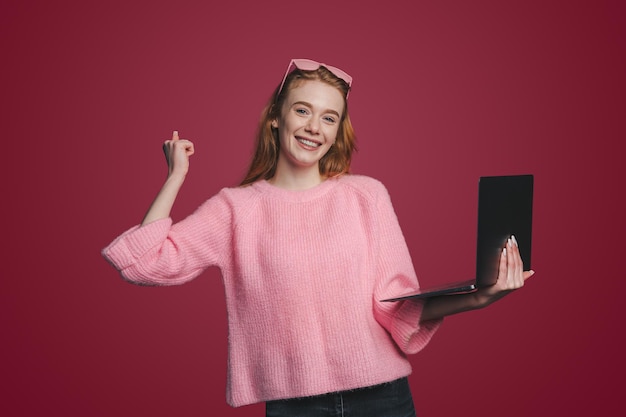 Photo portrait of attractive lovely redhaired woman holding in hands laptop celebrating winning isolated over red color background