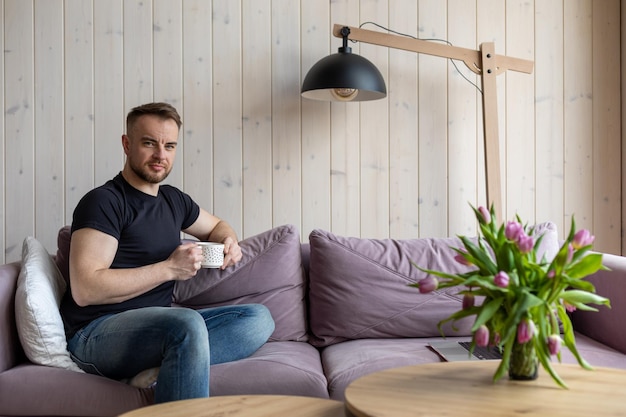 Photo portrait of attractive man holding white cup of coffee and sitting on soft violet sofa in casual clo