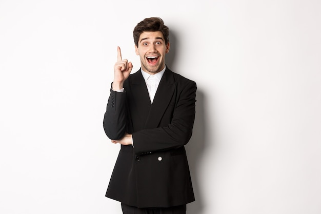 Portrait of attractive man in suit, having an idea, standing excited and raising one finger to tell suggestion, think-up solution, standing over white background