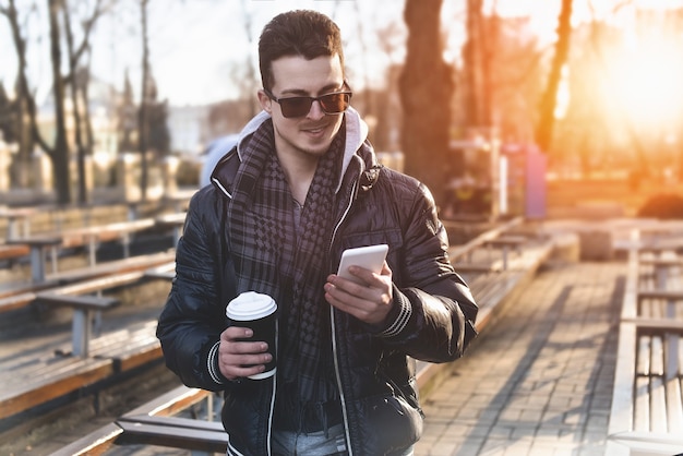 Портрет привлекательного мужчины в куртке, использующего мобильный телефон и держащего кофе на вынос