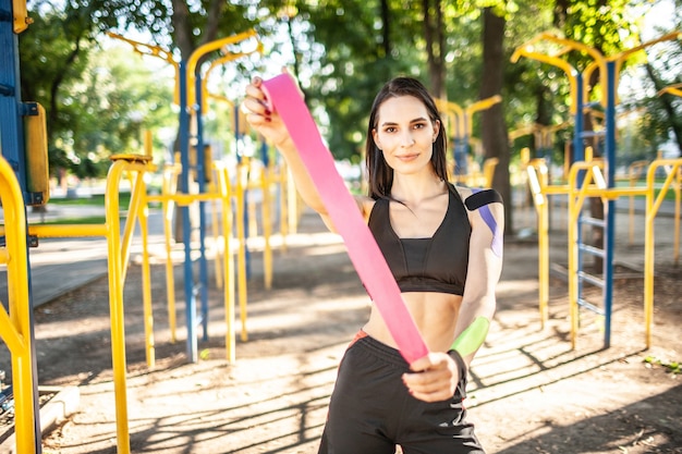 Portrait of attractive muscular brunette woman wearing black sports outfit, looking at camera. Young smiling female athlete posing with pink kinesio tape in hands. Concept of sports treatment.