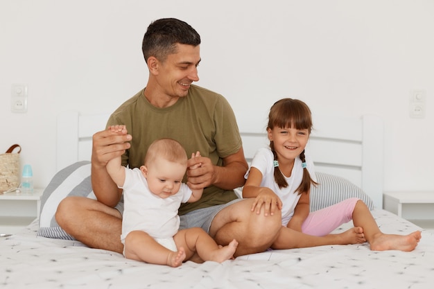 Portrait of attractive nan wearing green casual t shirt sitting on bed with his little children, holding infant baby hands, looking at cute elder daughter with smile and love.