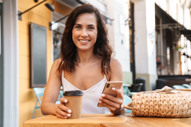 Portrait of attractive pleased woman with straw bag drinking coffee from paper cup and using cellphone while sitting in cozy cafe outdoors