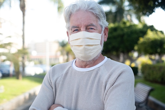 Portrait of attractive senior man standing outdoor in public park with crossed arms wearing mask