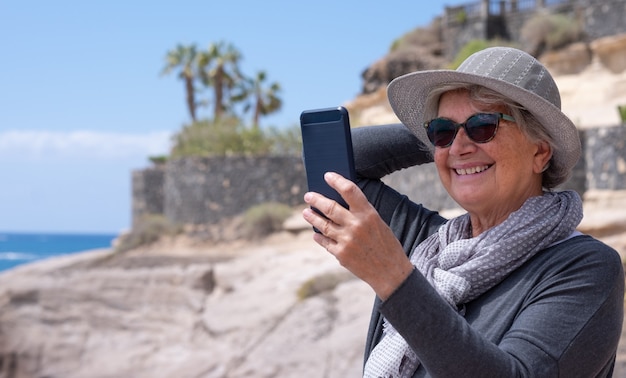 Ritratto di attraente donna anziana dai capelli bianchi utilizzando smartphone all'aperto al mare sorridendo felice. concetto di stili di vita gioiosi, pensionamento felice