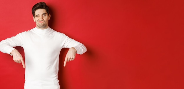 Portrait of attractive skeptical young man in white sweater grimacing and pointing fingers down disagree and dislike something standing over red background