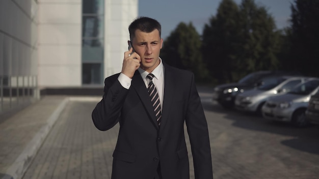 Портрет привлекательного стройного бизнесмена в строгом костюме, говорящего по смартфону на фоне бизнес-центра в теплый солнечный день