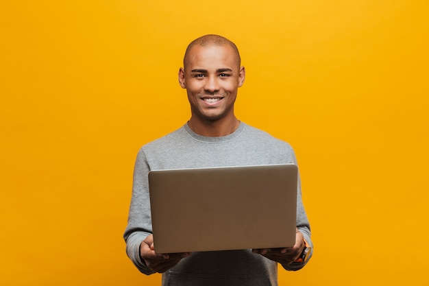 Photo portrait of an attractive smiling confident casual young african man standing over yellow wall, holding laptop computer