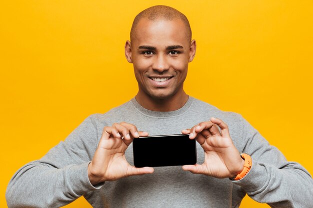 Ritratto di un giovane africano attraente e sicuro di sé, sorridente, in piedi sul muro giallo, che mostra un telefono cellulare con schermo vuoto