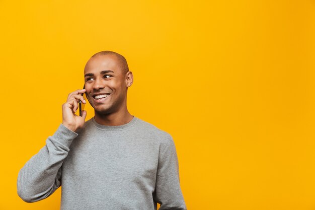 Ritratto di un giovane africano attraente e sicuro di sé sorridente in piedi sul muro giallo, parlando al cellulare