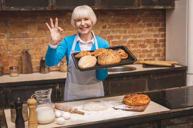 Portrait of attractive smiling happy senior aged woman is cooking on kitchen. Grandmother making tasty baking. Ok sign.