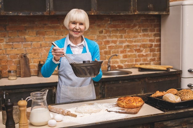 Il ritratto della donna invecchiata senior felice sorridente attraente sta cucinando sulla cucina. nonna che produce una cottura saporita.