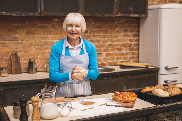 Portrait of attractive smiling happy senior aged woman is cooking on kitchen. Grandmother making tasty baking.