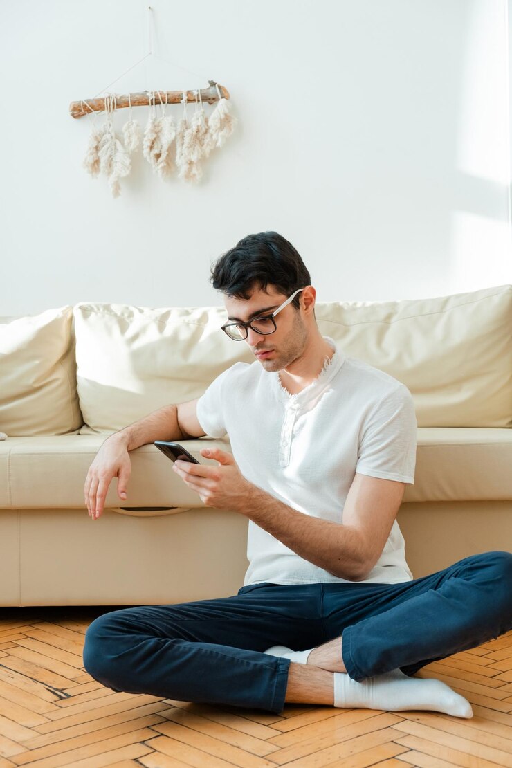 Portrait of an attractive smiling man in casual clothes sitting on the floor near the sofa in the living room and watching the news using a mobile phone