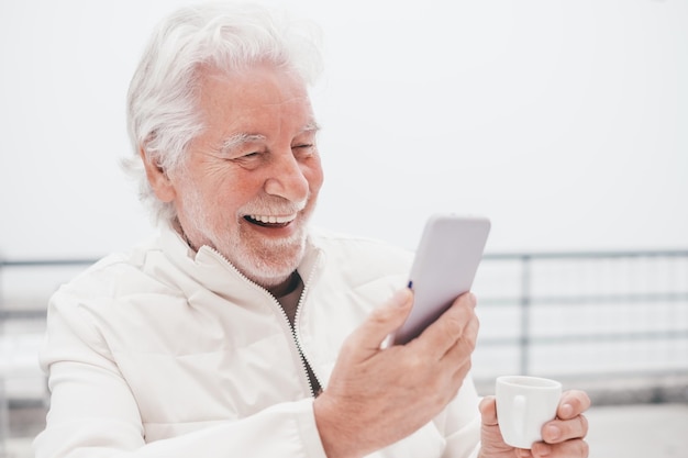 Portrait of attractive smiling senior man sitting outdoors at cafe table on a foggy day using phone